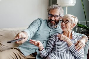 Un hombre y una mujer de mediana edad sentados cómodamente en un sofá. El hombre apunta con un control remoto.