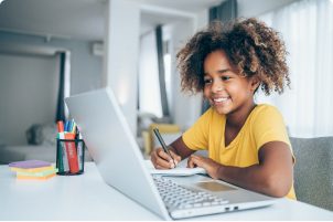 Una niña de 10 años aproximadamente mira su laptop y toma nota con un lápiz y papel.