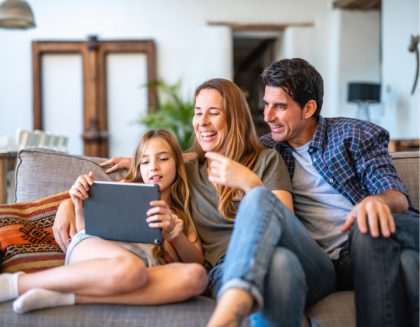 Una mamá, un papá y su hija están sentados en un sofá. Sonríen y miran la tablet que la hija está sosteniendo.