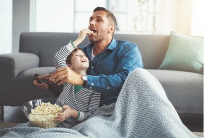 Un padre y su hijo disfrutando de una película mientras comen palomitas de maíz. El padre está sosteniendo un control remoto.