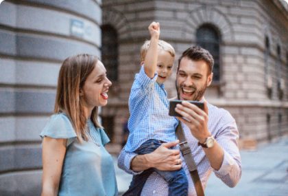 Una madre, un padre y su hijo pequeño fuera de un edificio en un día soleado. El padre sostiene un smartphone para que su hijo lo vea y todos vitorean.