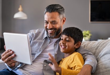 Un padre y un hijo sentados en un sofá, riéndose mientras miran contenido en una tableta.