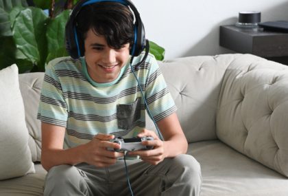 Un adolescente con camiseta a rayas, auriculares y micrófono sentado en un sofá blanco juega con un videojuego.