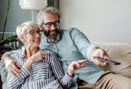Un hombre y una mujer de mediana edad sentados cómodamente en un sofá. El hombre apunta con un control remoto.