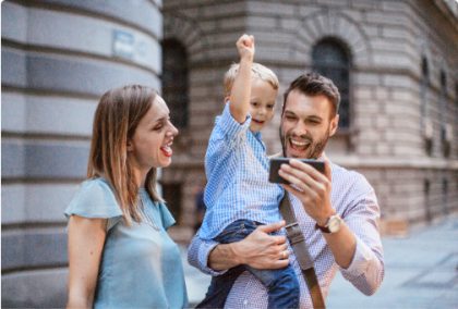 Una madre, un padre y su hijo pequeño fuera de un edificio en un día soleado. El padre sostiene un smartphone para que su hijo lo vea y todos vitorean.