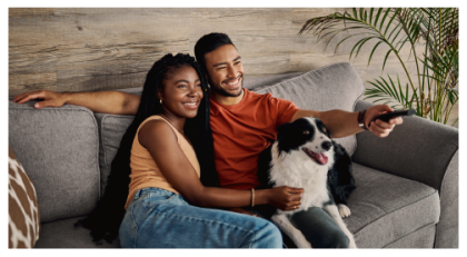 Pareja y perro sentados en el sofá sonriendo mientras sostienen el control remoto de la televisión