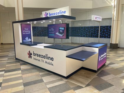 Un kiosco de Breezeline de color blanco y azul marino en un centro comercial, con pantallas digitales con promociones, sobre un piso de baldosas de varios colores.