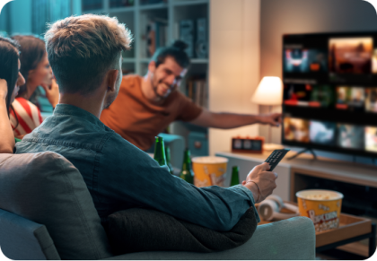 Un grupo de amigos se relaja en una sala con luz tenue. Una persona está sentada en un sofá, sostiene un control remoto y lo apunta hacia un TV donde se ve un menú de streaming.