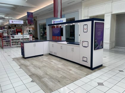 Un kiosco de Breezeline en forma de L, de color blanco y azul marino en un centro comercial, con pantallas digitales, gabinetes y una sección con piso símil madera.