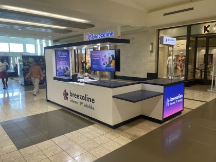 Interior de un centro comercial de varios niveles con tiendas, en donde aparece un kiosco de Breezeline.