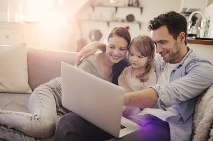 Una madre, un padre y su hija pequeña sonríen mientras miran la pantalla de una laptop sentados juntos en un sofá.