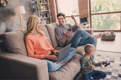A family relaxes in a living room using a laptop, smartphone, and gaming controller together.