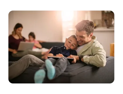 A father and young son smile while cuddling on a sofa and looking at a digital tablet.