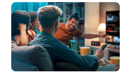 Grupo de amigos sentados en un sofá, mirando TV juntos, con bocadillos y bebidas sobre la mesa en una sala acogedora.