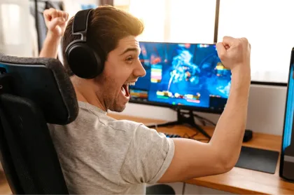 An excited man wearing headphones cheers with raised fists while playing a video game on a computer.