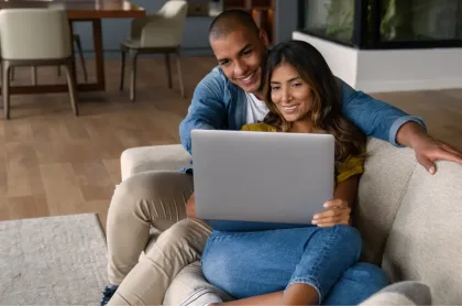 A smiling couple sits closely together on a couch while looking at a laptop computer.