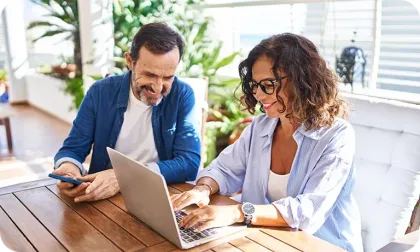 A smiling man and woman sit at an outdoor wooden table together, looking at a laptop and a smartphone.