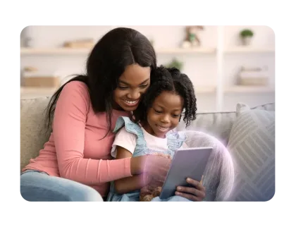 A mother and her young daughter smile while using a tablet together in a bright living room.