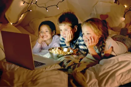 Three children lie on a bed with popcorn, happily watching a movie on a laptop under fairy lights.