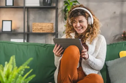 Una mujer de cabello rizado con auriculares se ríe mientras mira una tablet en un sofá verde.