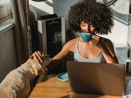 Mujer bebiendo sorbos de una taza en una mesa con una laptop mientras acaricia a un perro
