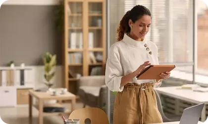 A woman in a white blouse and tan trousers uses a tablet while standing in a bright, modern office space.