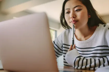 A young woman thoughtfully holds a pen to her chin while looking at her laptop screen.