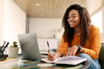 A smiling woman with curly hair takes notes in a journal while looking at a laptop in a bright room.